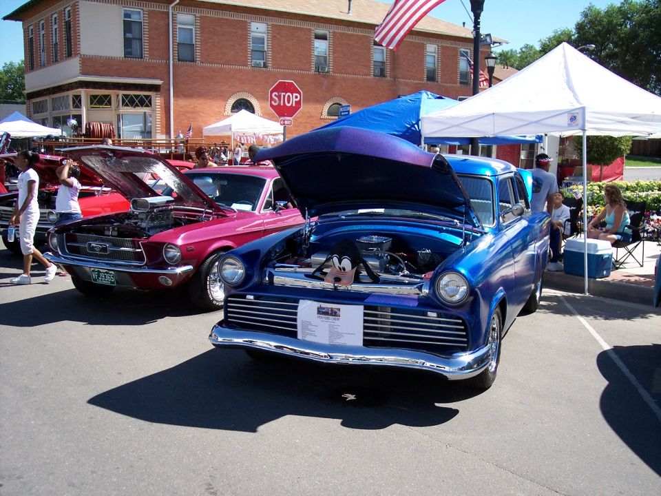 Blue classic car with hood open, showing engine. Pink Ford Mustang also visible with hood open. Outdoor car show setting