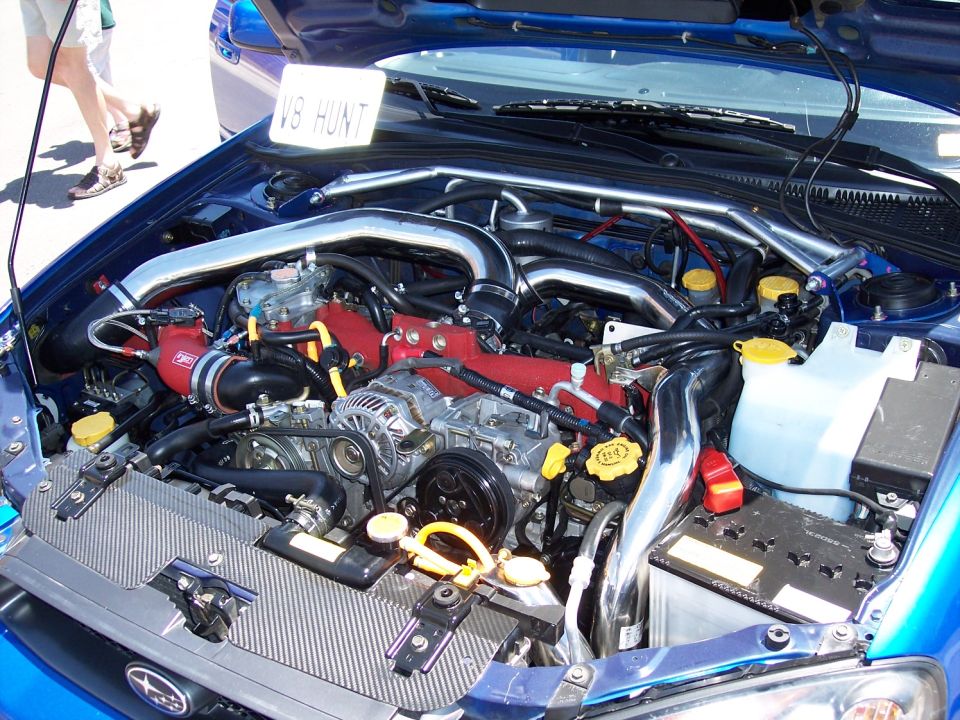 Blue Subaru engine bay with chrome piping and red engine components. A sign reads "V8 HUNT"