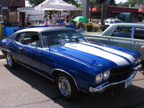 1989 Ford Mustang - A blue 1970 Chevrolet Chevelle hardtop with white hood and deck stripes and a black vinyl roof sits at a downtown street show, viewed from…
