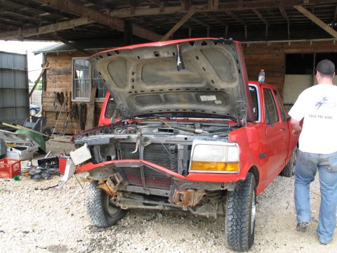 1995 Ford Bronco - A red truck with its hood open and a damaged front end is parked under a wooden shelter.