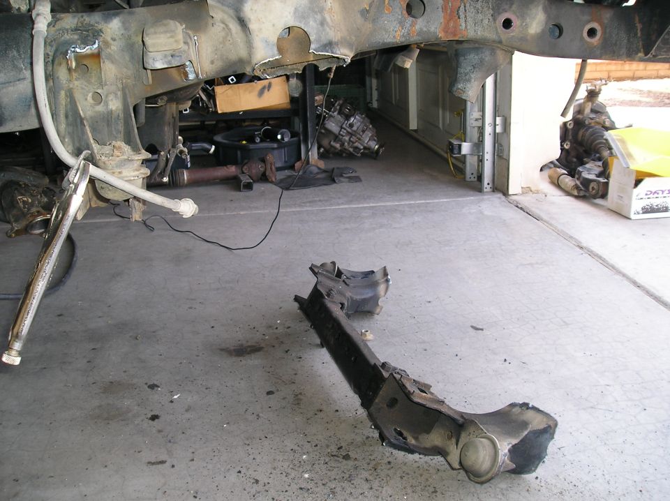 Vehicle frame and suspension components on a concrete floor, some parts appear rusted and disassembled