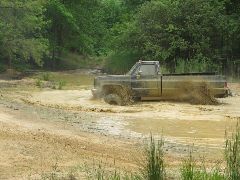 1976 Chevrolet Blazer - A pickup truck navigates a muddy off-road course, kicking up water and dirt.