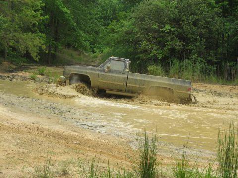 1976 Chevrolet Blazer - A muddy pickup truck navigates through a water-filled off-road course, kicking up spray.