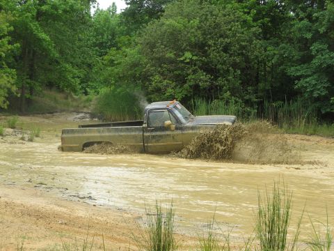 1976 Chevrolet Blazer - A truck is stuck in the mud, creating a large splash as it tries to move through the water.