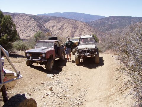 1967 Ford Bronco - Several off-road vehicles are stopped on a dusty mountain trail under a clear blue sky.