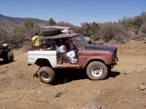 1967 Ford Bronco - Two off-road vehicles navigate a dusty trail in a dry, hilly landscape under a clear blue sky.