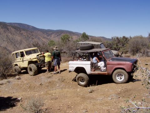 1967 Ford Bronco - A red and white Ford Bronco is parked on a dirt road with another yellow vehicle nearby, under a clear blue sky.