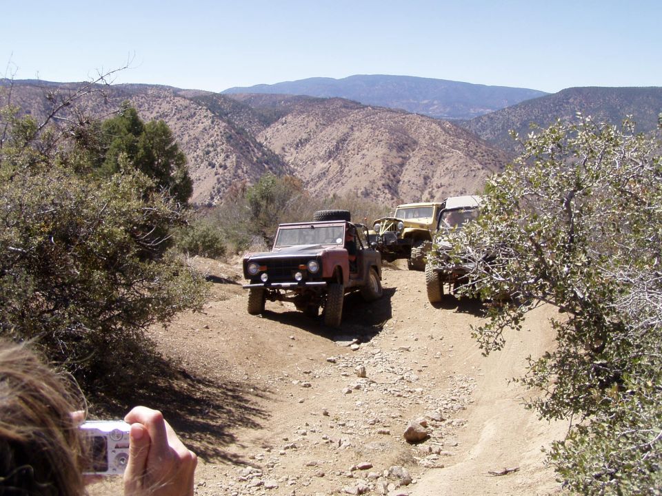 Several off-road vehicles, including a red early Ford Bronco, drive up a dusty mountain trail