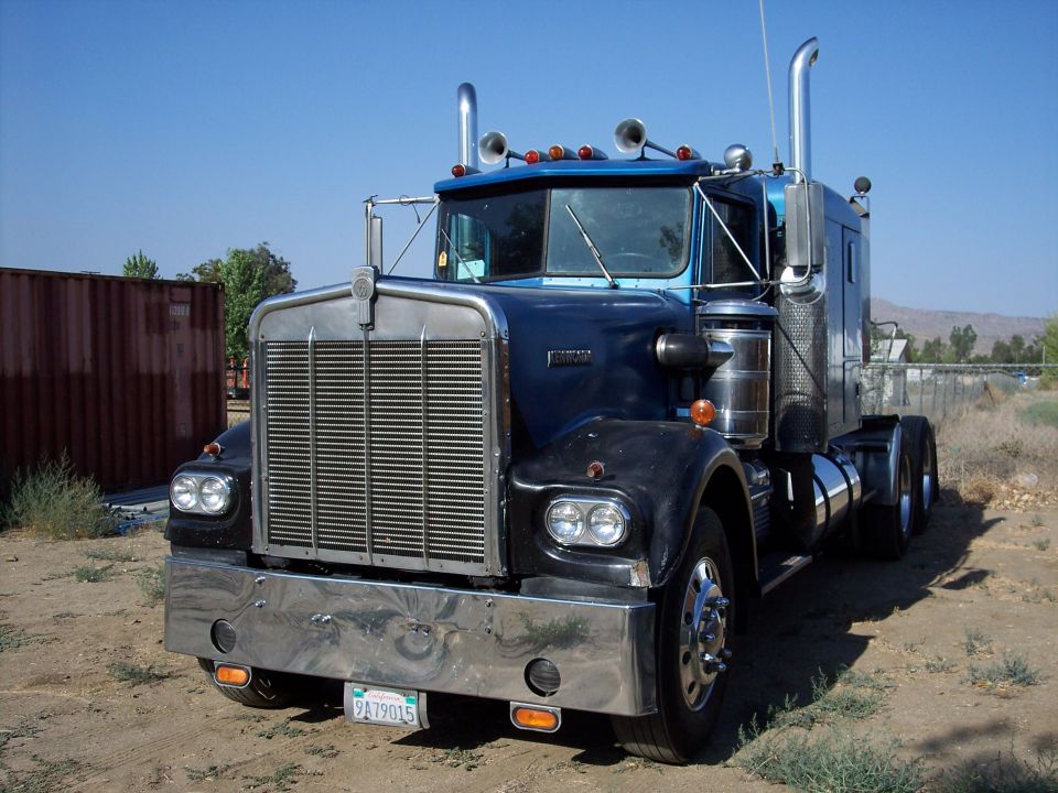 Dark blue Kenworth semi-truck with chrome grille and dual stacks on a dirt lot
