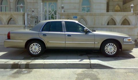 2007 Mercury Grand Marquis - A tan 2007 Mercury Grand Marquis is parked on a curb, its side profile against an ornate building.