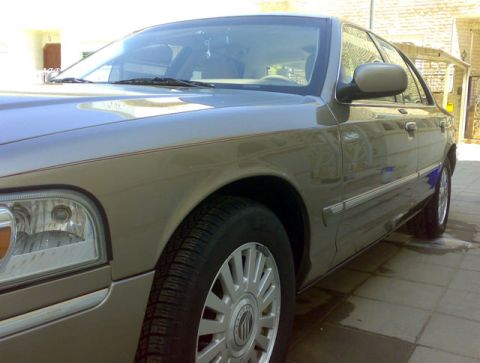 2007 Mercury Grand Marquis - A tan Mercury Grand Marquis is viewed from the front passenger side at a low angle, showing its chrome trim and alloy wheels.