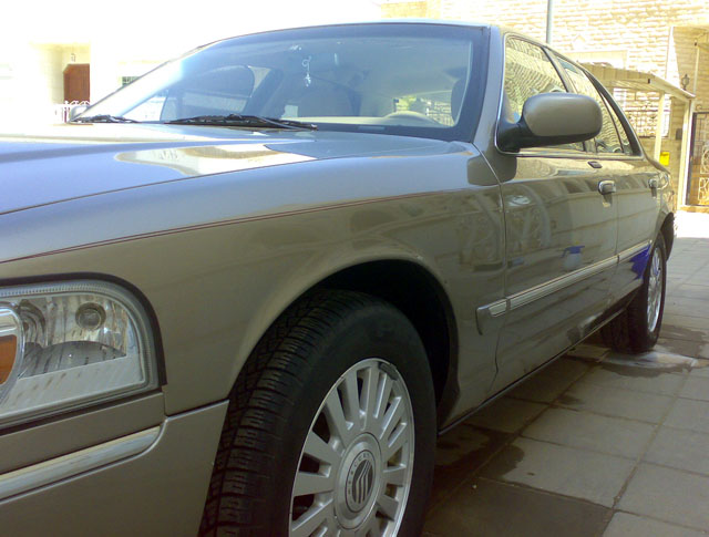 Tan Mercury Grand Marquis, front passenger side, low angle. Chrome trim along the lower body and door handles. Alloy wheels visible