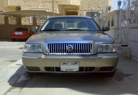 2007 Mercury Grand Marquis - A gold Mercury Grand Marquis is parked in front of a stone house, with another red car visible in the background.