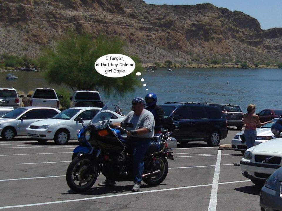 Two people on a blue motorcycle parked near a lake, with cars in the background