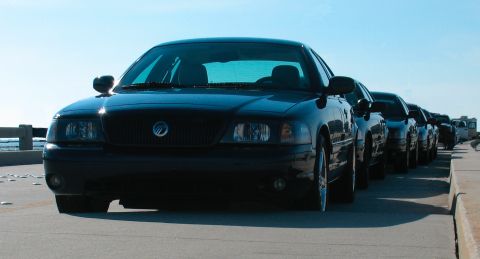 1997 Ford Crown Victoria - A line of black Mercury Marauders are parked along a roadside with the ocean in the background.