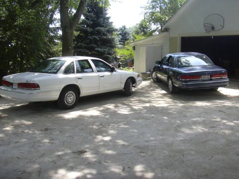 1997 Ford Crown Victoria - Two Ford Crown Victorias are parked in a driveway next to a garage.