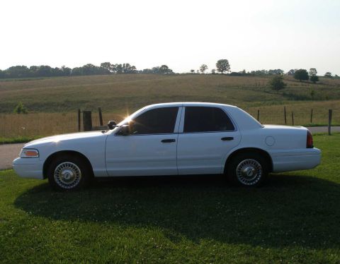 2001 Ford Crown Victoria - A white 2001 Ford Crown Victoria is parked on a grassy area with a fence and rolling hills in the background.