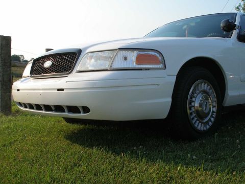2001 Ford Crown Victoria - The front of a white 2001 Ford Crown Victoria is visible, parked on a grassy area with a wooden post to the left.