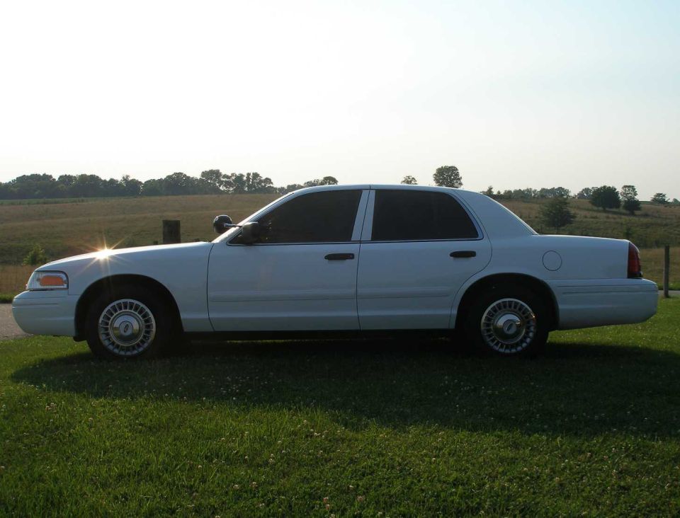White sedan parked on grass with a field and trees in the background