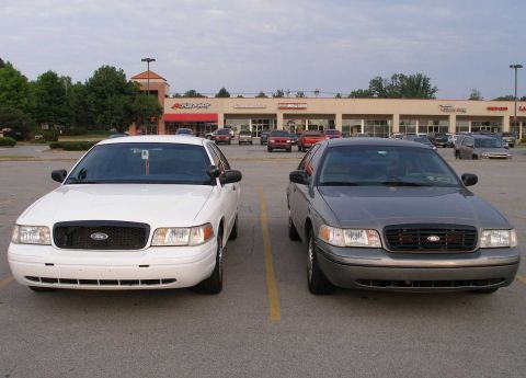 2001 Ford Crown Victoria - Two Ford Crown Victorias are parked in a lot in front of a strip mall.
