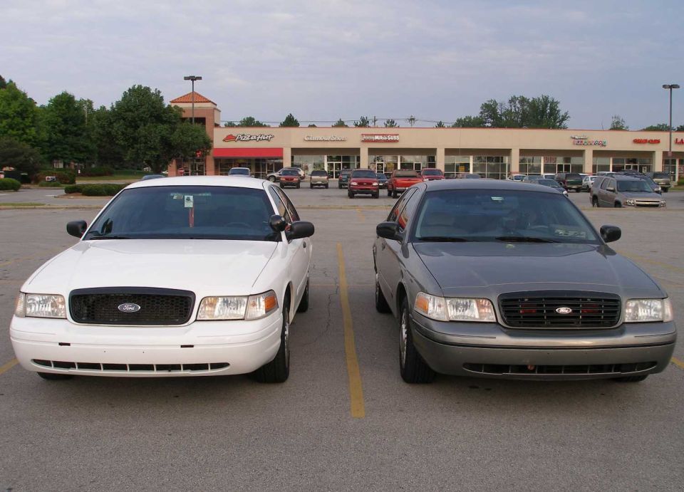 Two Ford Crown Victorias parked side-by-side, one white, one gray. Front view