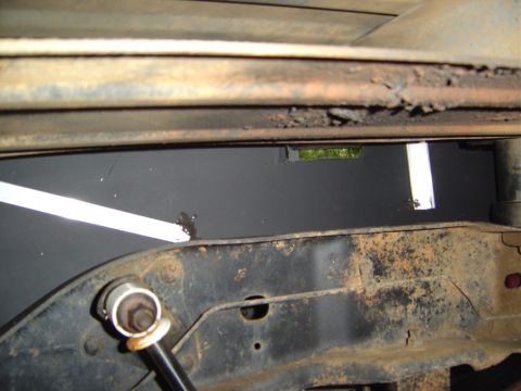 1986 Ford Bronco - Close-up looking through a rusty body seam at the backside of a freshly painted black panel bolted behind the frame rail, with light leakin…