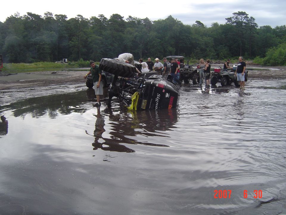 ATV flipped over in shallow water with people around it. Other ATVs are parked nearby