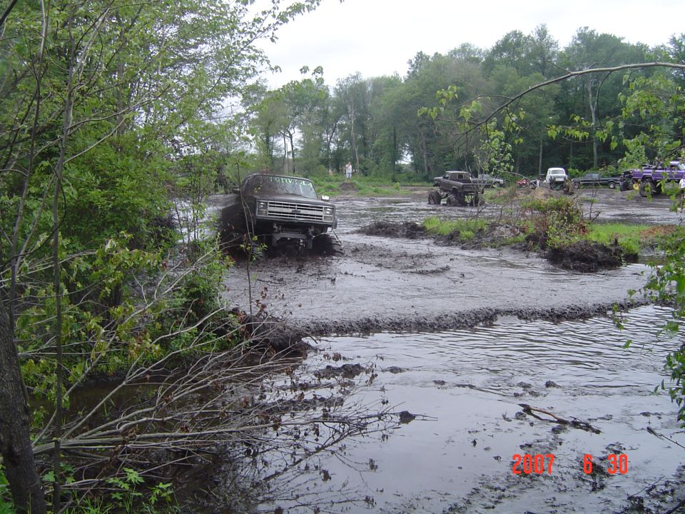 Muddy trucks driving through a swampy area. Other vehicles are visible in the background