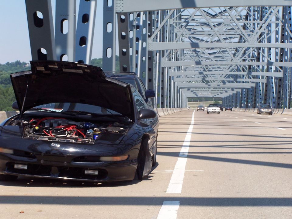black 1994 Ford Probe with hood open on a bridge, other cars on the road