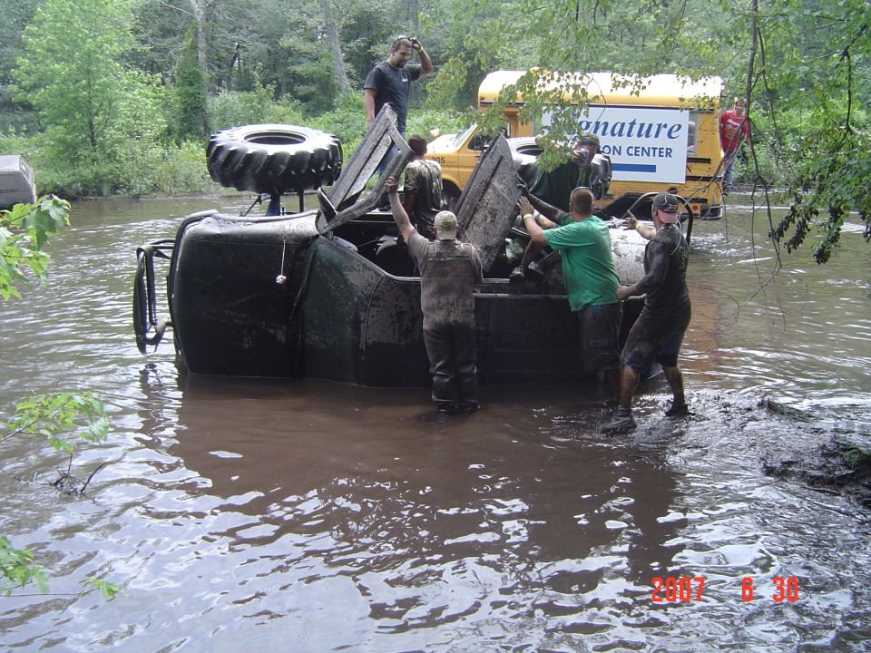 Muddy off-road vehicle on its side in water, with people attempting to right it. A yellow bus is in the background