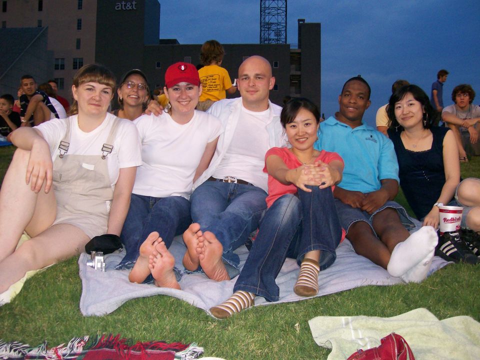 Group of people sitting on a blanket on grass, with buildings and a tower in the background