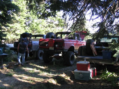 1979 Ford Bronco - Multiple modified off-road vehicles are gathered in a wooded area, with people attending to one of them.