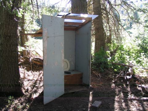 1979 Ford Bronco - An old, weathered outdoor toilet stands open in a wooded area, with sunlight filtering through the trees.
