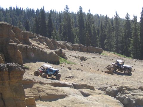 1979 Ford Bronco - Off-road vehicles traverse a challenging rocky landscape with a dense forest in the background.