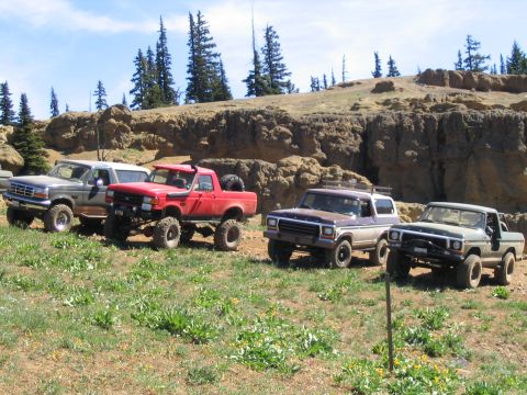 1979 Ford Bronco - Four modified off-road vehicles, including a 1979 Ford Bronco, are parked on a grassy slope with rocky terrain in the background.