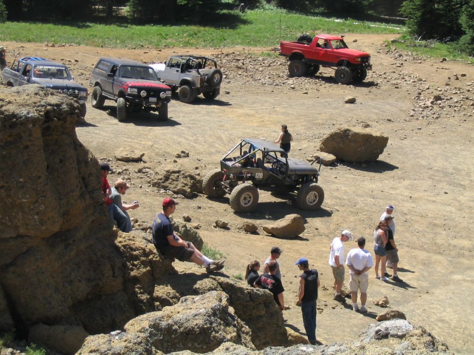 Several off-road vehicles, including a red pickup truck and a buggy, are parked on a dirt trail with people standing around