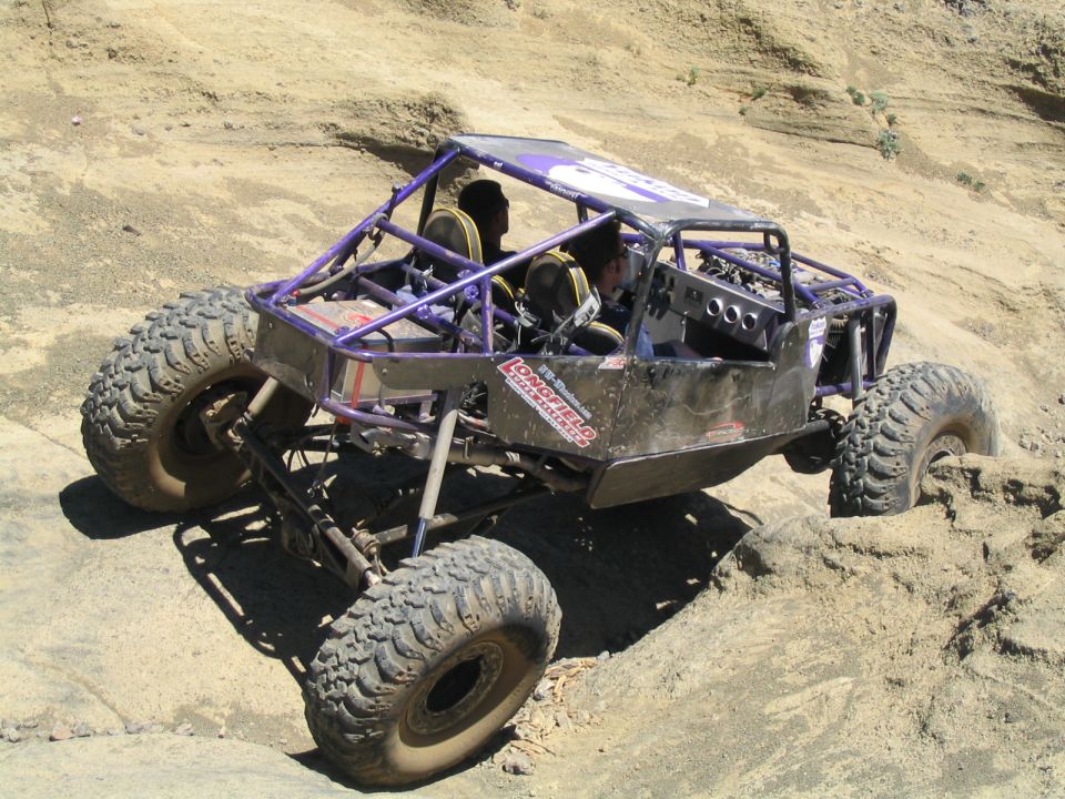Custom rock crawler buggy with a roll cage, large tires, and two occupants, navigating sandy terrain