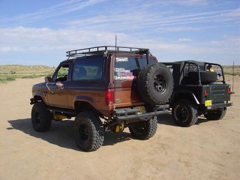 1986 Ford Bronco II - A brown 1986 Ford Bronco II is parked on sandy terrain, with a green Jeep visible to its right. Both vehicles appear to be modified for off…