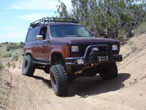1986 Ford Bronco II - Modified 1986 Ford Bronco II navigates an off-road trail with large tires and a winch.