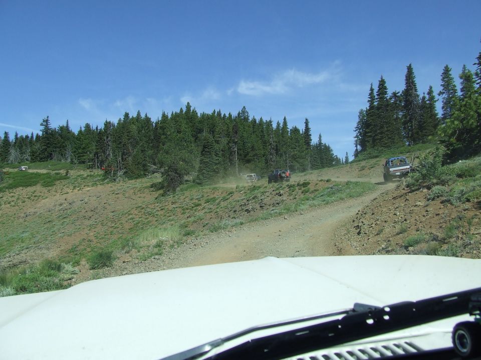 White vehicle hood in foreground, dirt road with multiple SUVs driving on a forested hillside under a blue sky