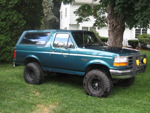 1996 Ford Bronco - A teal 1996 Ford Bronco with a suspension lift and off-road tires sits on a grassy lawn in front of a white house.