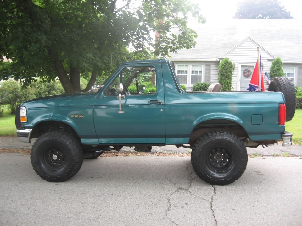 Teal-green lifted Ford Bronco, top removed, side view on suburban street with mud tires