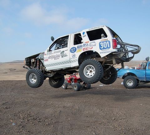 1992 Ford Explorer - A modified 1992 Ford Explorer is airborne during a race, with a blue truck and a red ATV nearby on a dirt track.