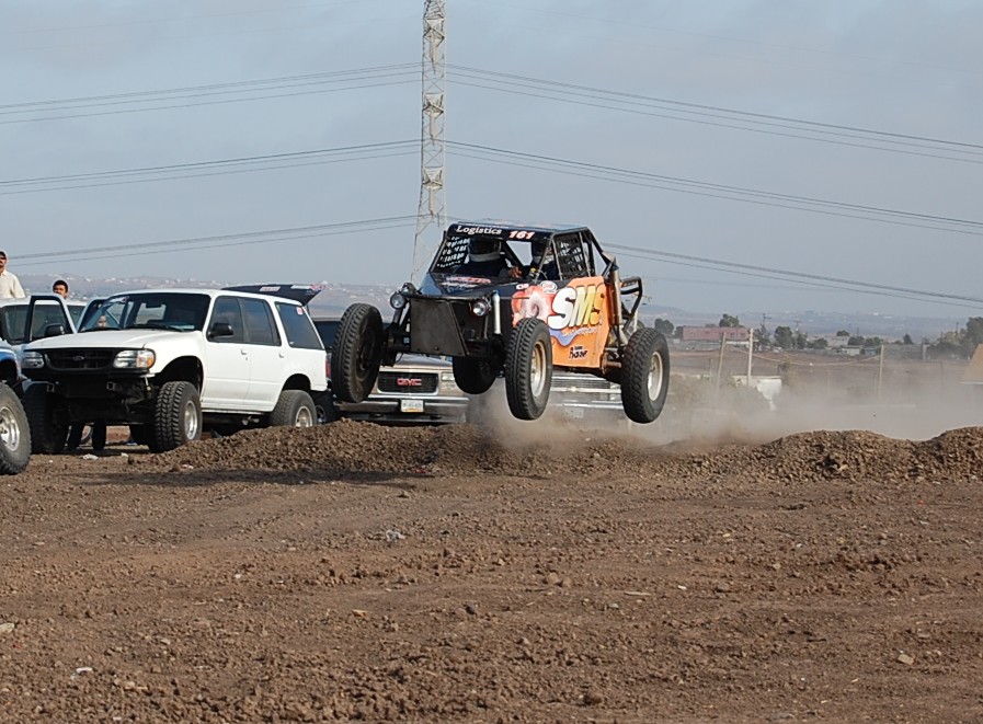 Buggy mid-air over dirt mound, white SUV and GMC truck nearby
