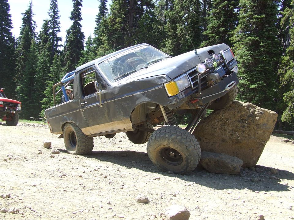 Dark gray SUV with large tires climbing over a rock, front right tire off the ground. Another red SUV is visible to the left