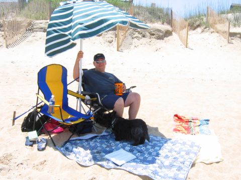 1985 Ford LTD Crown Victoria - A man and his dog enjoy a sunny day on the beach, sitting under a striped umbrella.