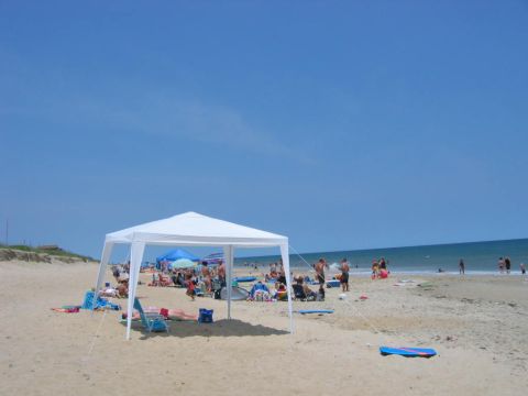 1985 Ford LTD Crown Victoria - A sunny day at the beach with many people enjoying the sand and surf under a large white canopy.