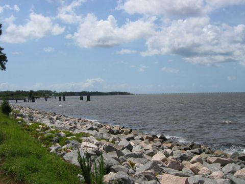 1985 Ford LTD Crown Victoria - A waterfront scene shows a rocky shoreline with grass and a distant bridge stretching across the water.