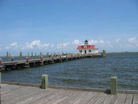 1985 Ford LTD Crown Victoria - A lighthouse with a red roof stands on a pier extending into the water under a blue sky with scattered clouds.