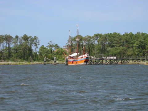 1985 Ford LTD Crown Victoria - A tall ship is docked at a pier on a body of water with trees lining the shore.